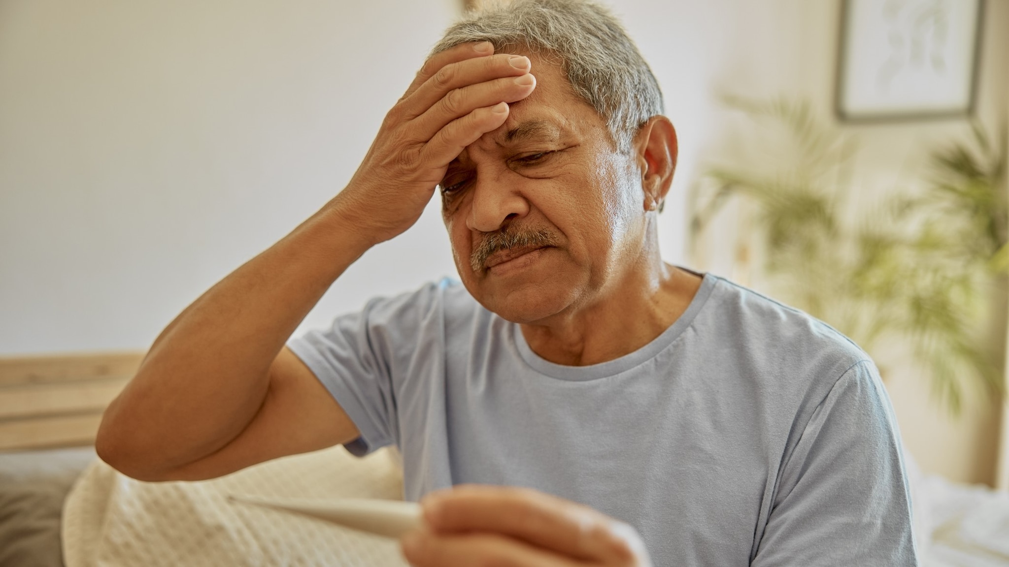 A person sitting with their hand on their head indicating a fever and looking at a thermometer.