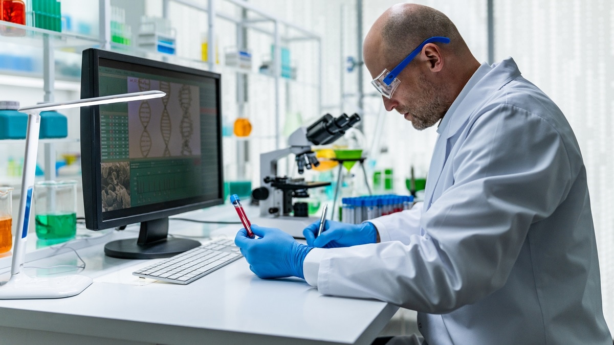 A male scientist in a white lab coat examines a blood sample at a laboratory