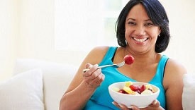 Overweight Woman Sitting On Sofa Eating Bowl Of Fresh Fruit