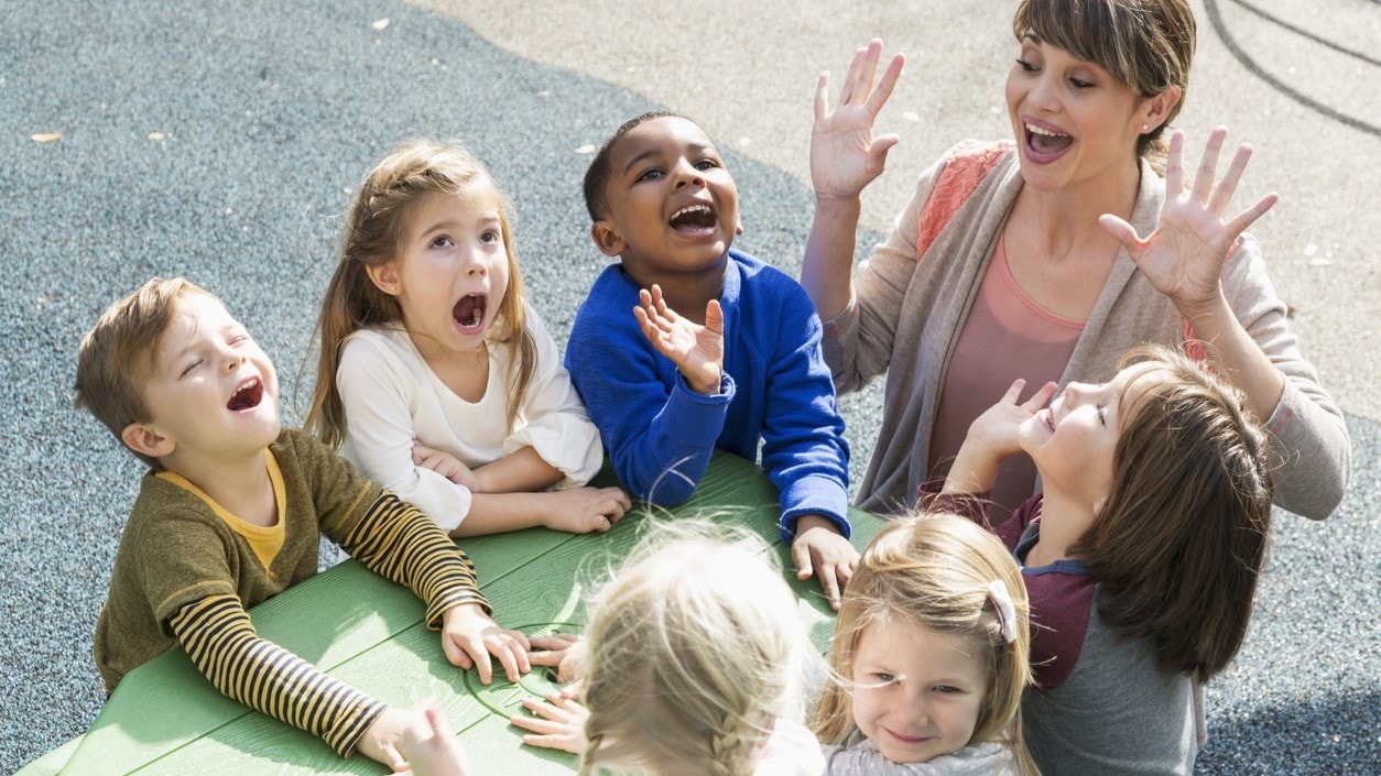 Teacher playing game outside at a table with six young children.