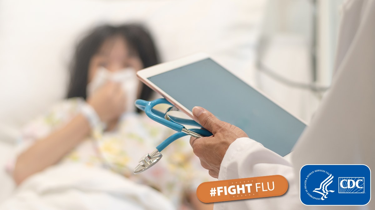 A doctor holding a tablet and a patient in a hospital bed.