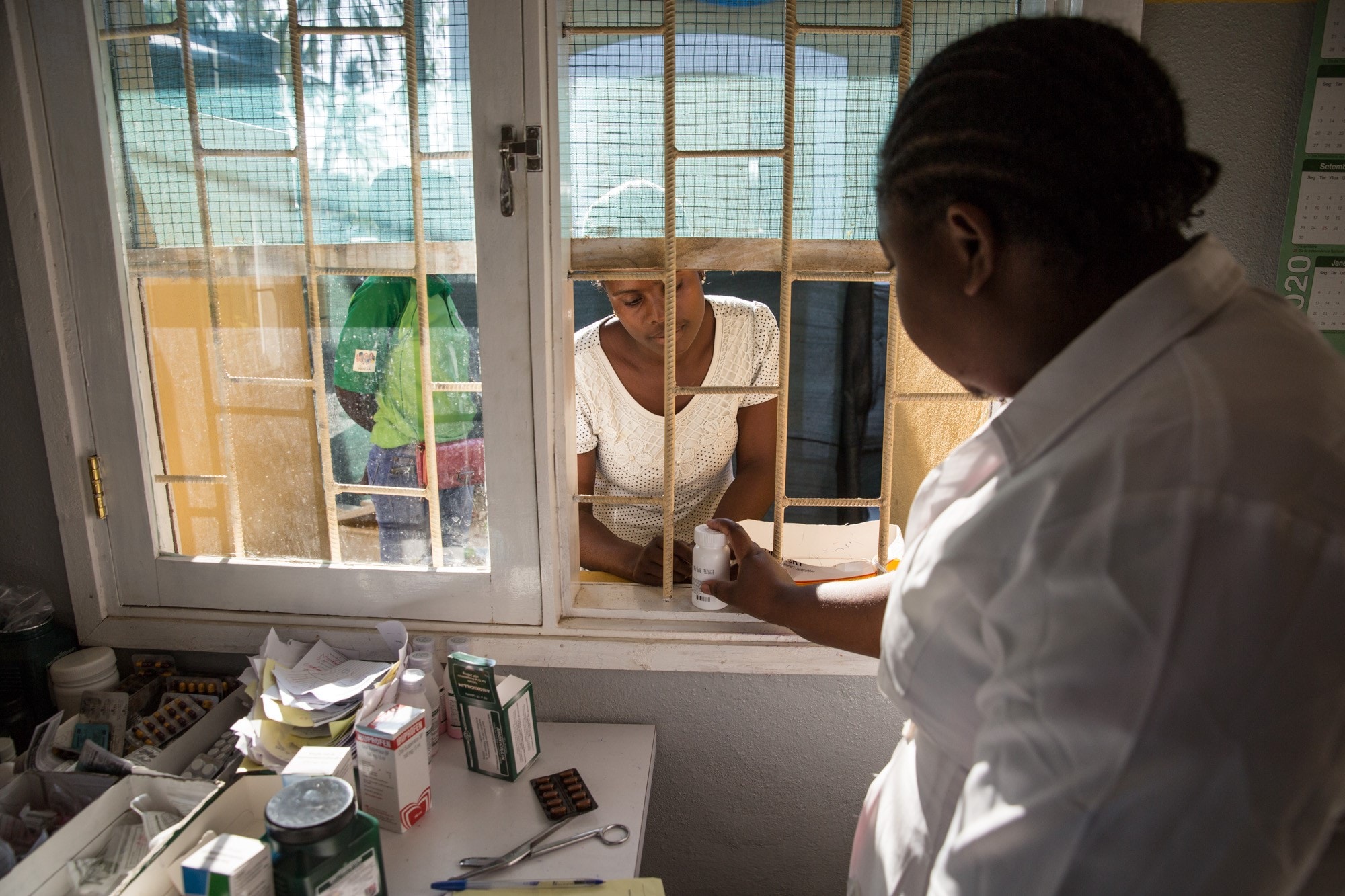 A woman in a white coat hands medication out a window.