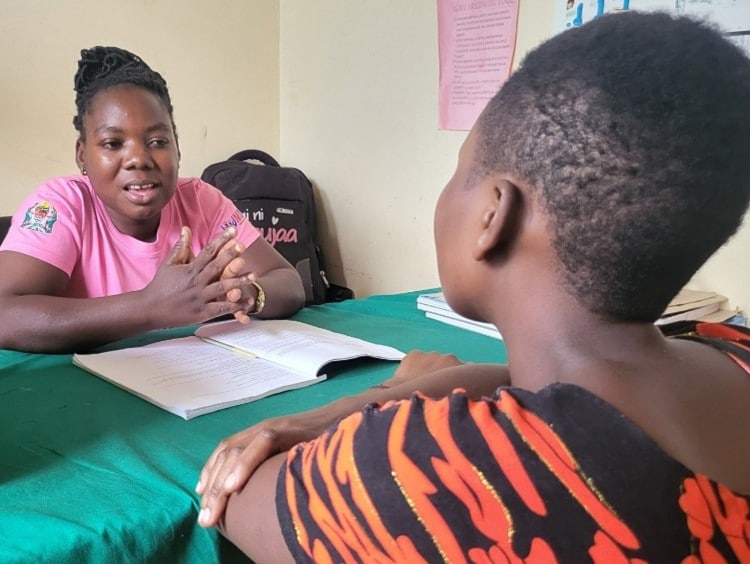 A young woman in a pink shirt speaks at a table with another woman.