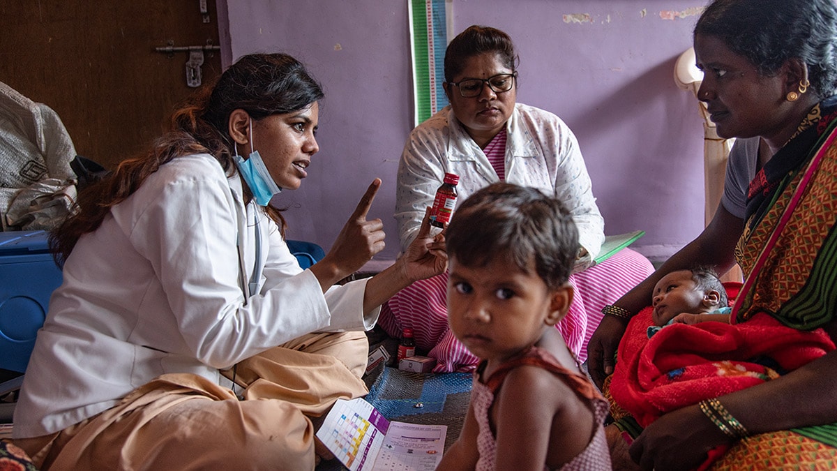 A mother with an infant and small child watches as 2 female healthcare workers conduct a routine immunization session.