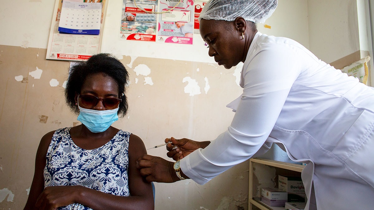 A pregnant woman sits in a chair while a health worker holds a syringe near her left arm.