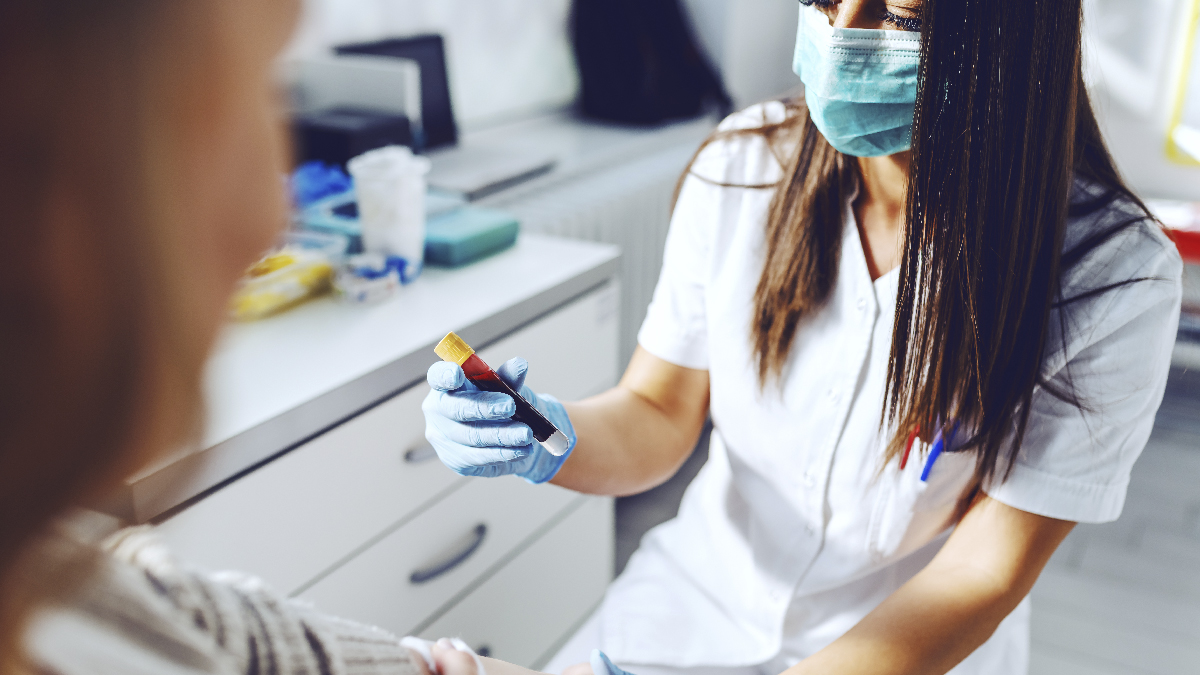 Woman in a medical setting, wearing a white medical uniform and a mask, holding a vial of blood.