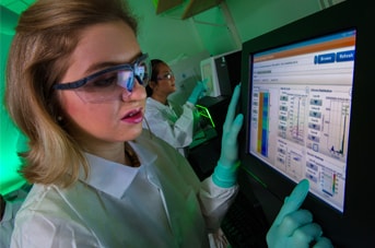 A woman with short blonde hair wears safety goggles and a lab coat while looking at lab results on a screen