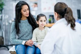 A woman and a child sit on a doctor's examine table in front of a doctor.