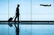 Silhouette of a traveler in an airport, walking with a rolling suitcase behind them as a plane takes off in the background.