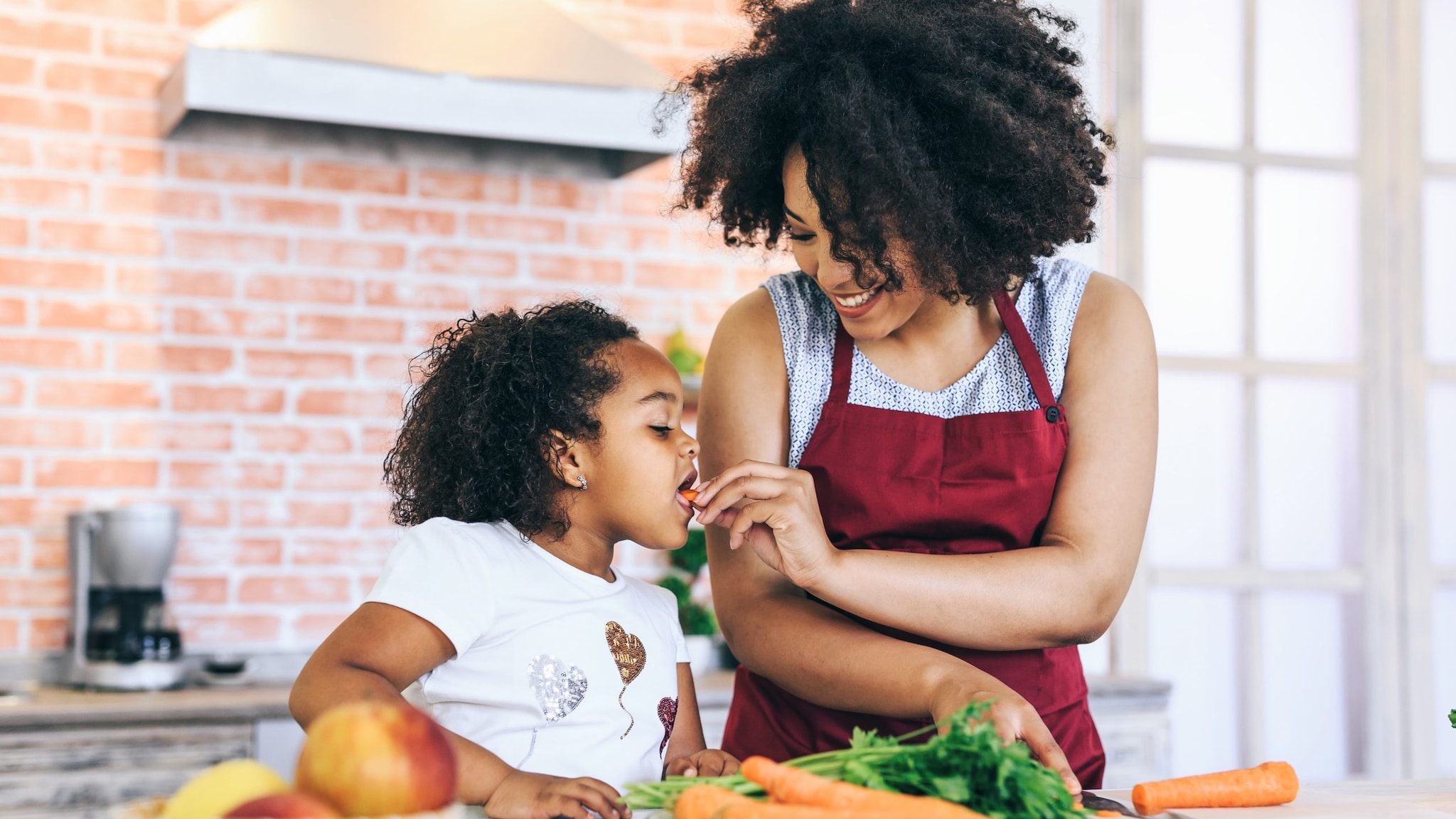 A picture of a mother and child in the kitchen together chopping vegetables. The child is eating a slice of carrot.