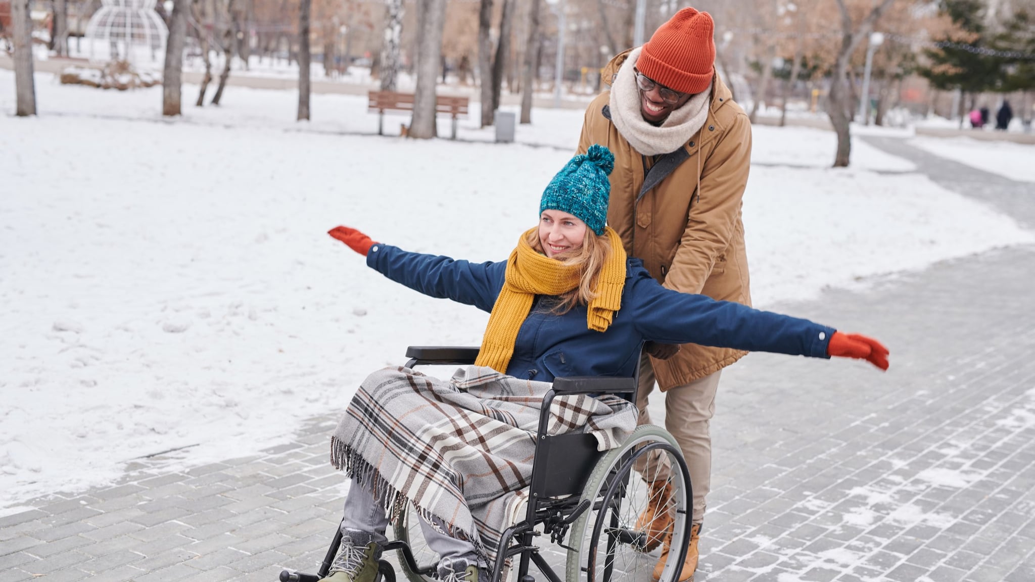 Two people in a snowy park. One is pushing the other in a wheelchair.