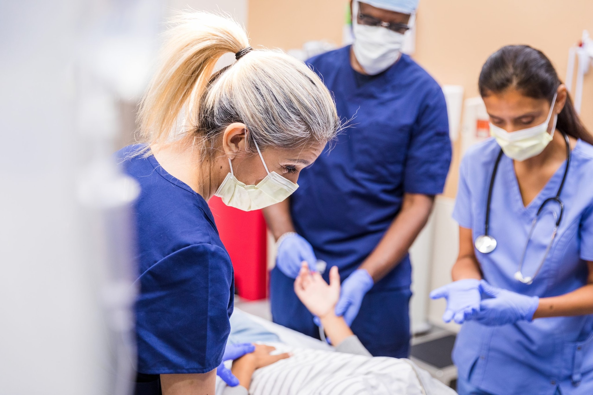 A three-person healthcare team stands around the bed of a patient as they administer care