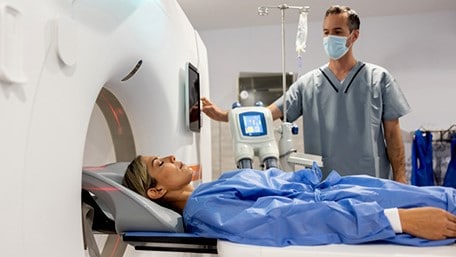 A woman lays on an exam table with her eyes closed as a health care professional prepares to send her through an imaging machine