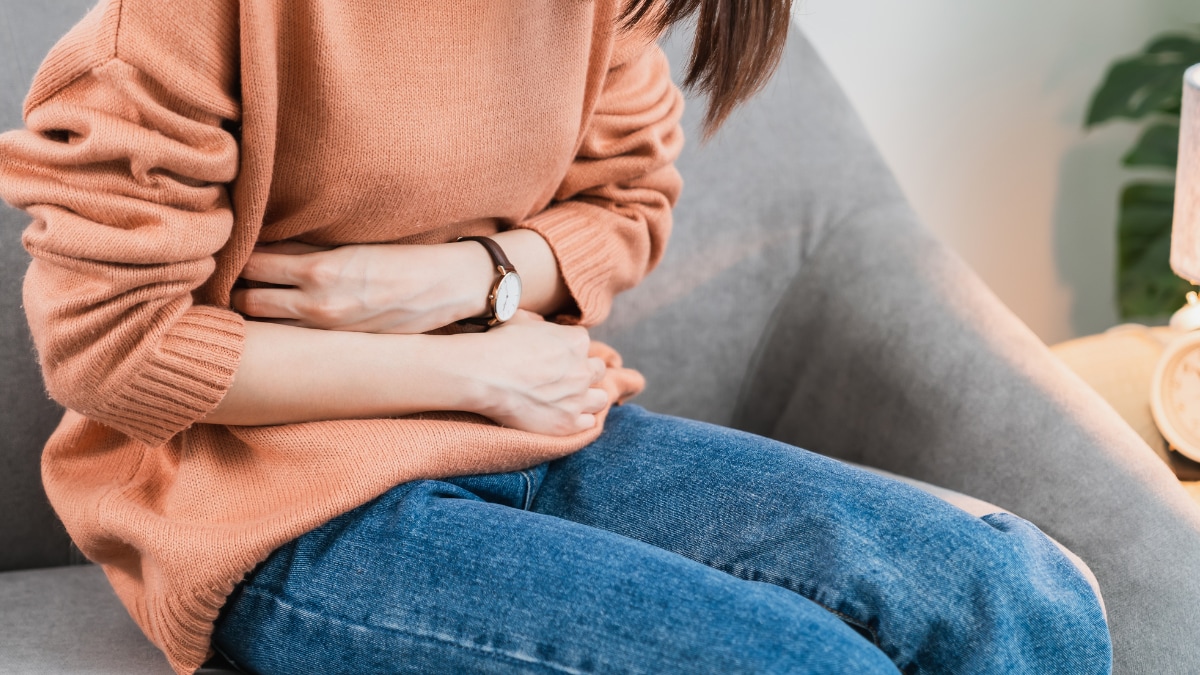 A person sitting down hunched over on a gray chair, holding their stomach with both hands.