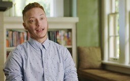 Hispanic transman sitting in room with bookcase behind him talking to camera