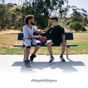 Image displays two athletic young men talking to each other while sitting on a bench on the side ostensibly on the side of a basketball court.