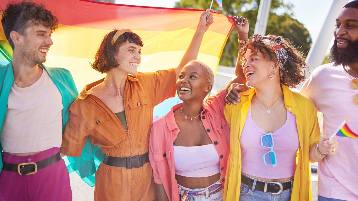 Hombres y mujeres jóvenes vistiendo una variedad de colores brillantes sosteniendo una bandera del orgullo arcoíris