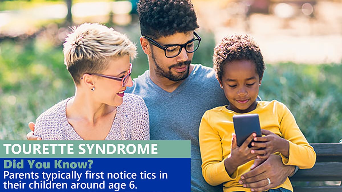 Parents sit on park bench with their daughter, looking and smiling at a cell phone.