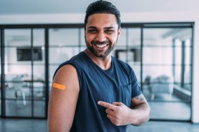 An adult man smiling and pointing to his arm after getting his shot.