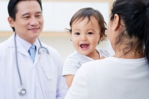 A woman with dark hair is holding a smiling baby and is talking to a male doctor.