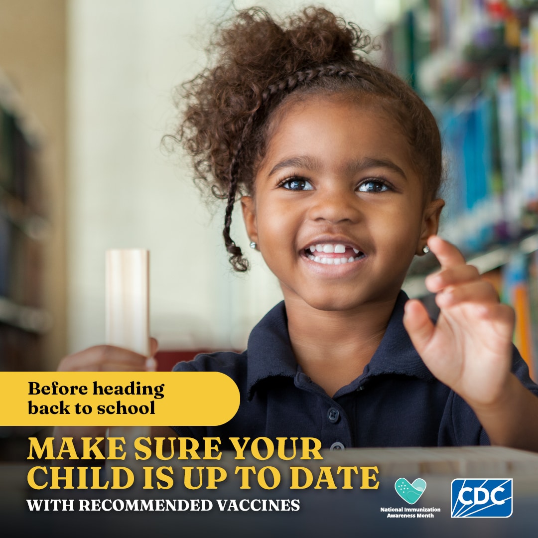 A little girl in a dark blue polo is holding a ruler in one hand and raises the other as she sits in a school library