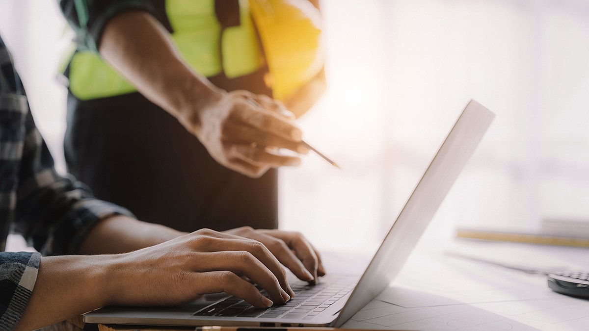 User imputs data via laptop and worker in construction gear points to screen with pencil.