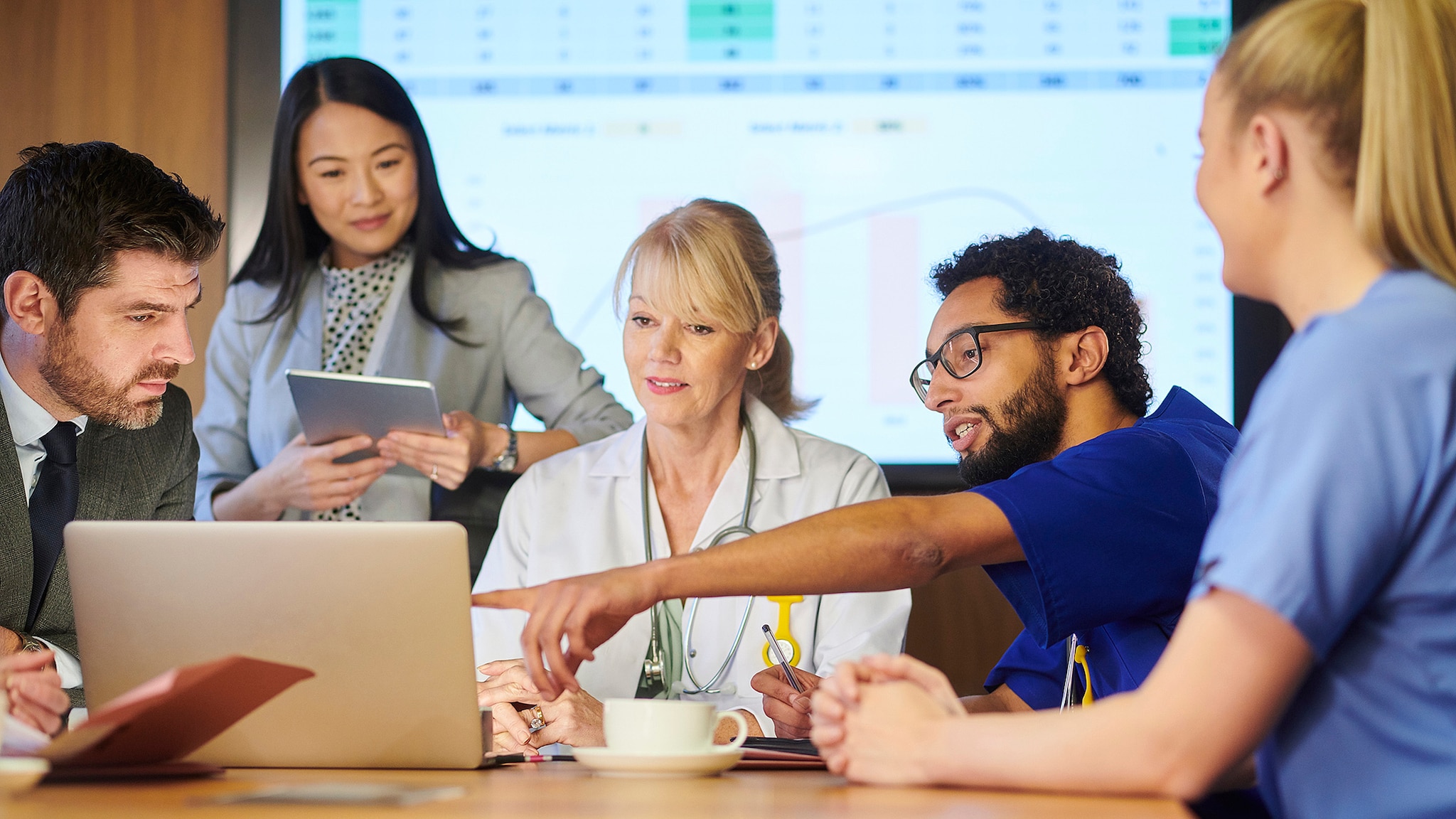 Group of business colleagues and medical professional going over health data on a laptop with charts on projected screen.